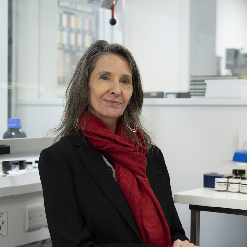 White woman with medium length dark hair sitting in a lab wearing a black jacket and red scarf