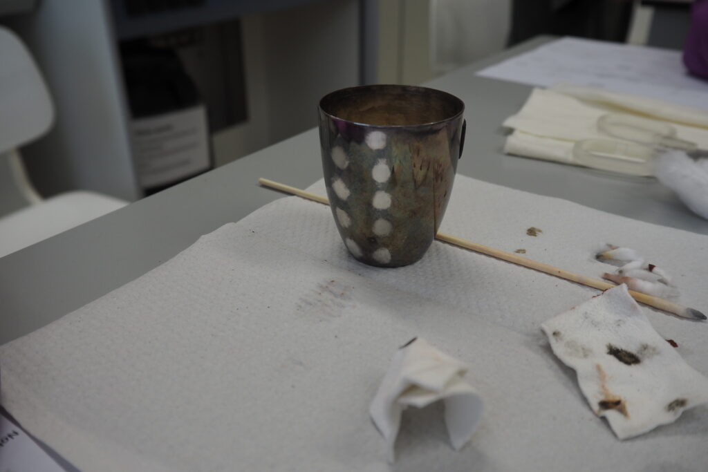 A heavily tarnished silver cup is placed on a paper towel. It has nine small patches of cleaning tests where the tarnish has been removed, and the lighter colour of silver is clearly visible. Some used cotton swabs lie next to the cup.